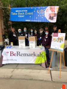 Photo of volunteers wearing standing behind a table underneath a banner announcing their Medication Take Back Event.